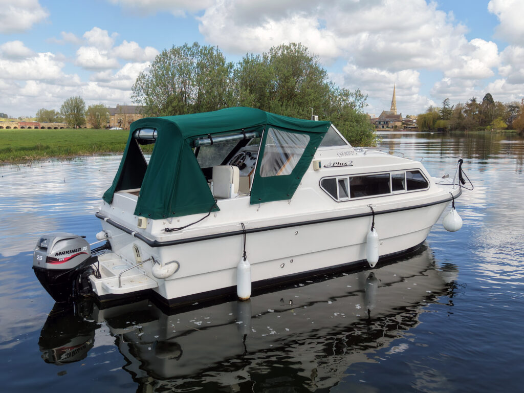 A white motorboat with a green canopy is anchored on calm water. Nearby greenery is visible, along with buildings in the background, including a church steeple. The boat features fenders and a small outboard motor.