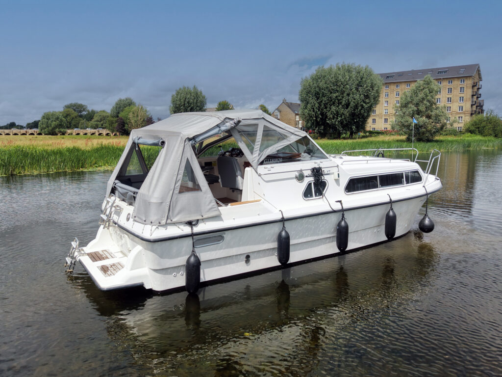 A white motorboat with a gray canopy is moored on calm water, surrounded by green vegetation. In the background, a large building is visible next to a row of trees under a clear blue sky.