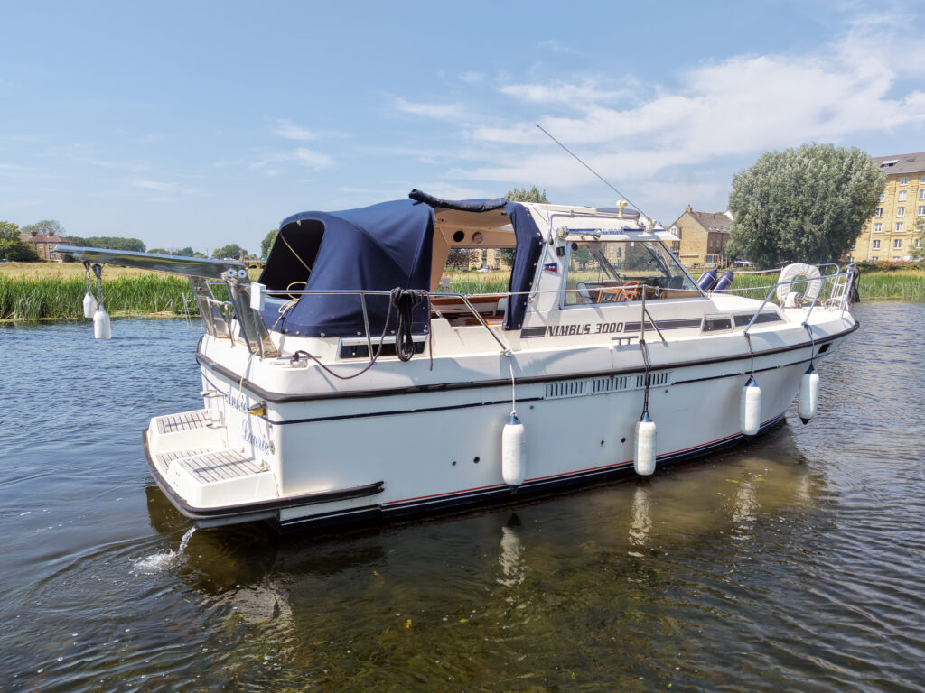 A white cruising boat with a blue canopy docked in calm water. The boat features railings and fenders, with green trees and buildings visible in the background under a clear blue sky.