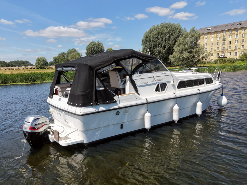 A white cabin cruiser boat with a black canopy is moored on a calm river. The background features lush greenery, and a large building with yellow siding is visible on the bank.