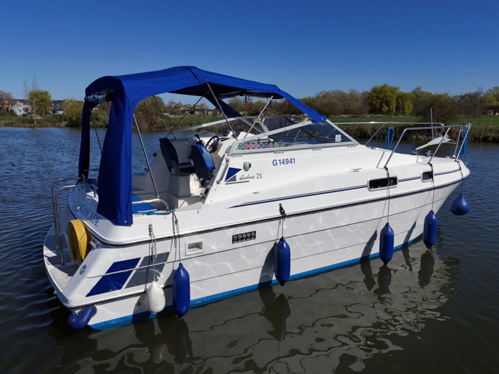 A white cabin cruiser boat with a blue canopy is docked on calm waters. The boat features blue fenders, a steering console located inside, and is surrounded by greenery in the background under a clear blue sky.
