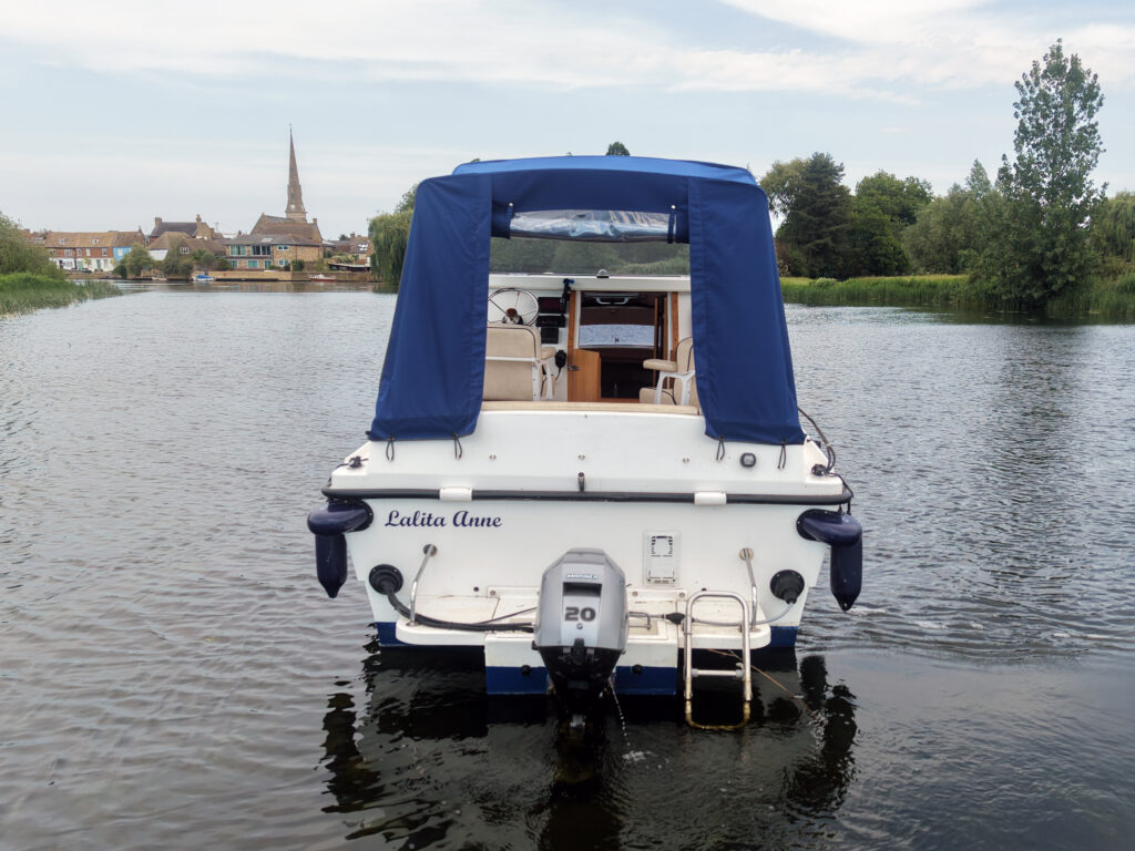 A white motorboat named "Ladita Anne" is docked on calm water, facing the viewer. The boat has a blue canopy and a motor labeled "20" attached to the rear. In the background, a small town is visible with a prominent church spire among trees.