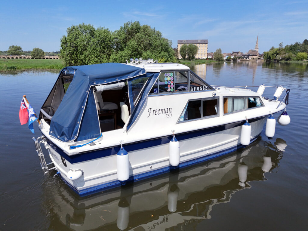 A white and blue Freeman 750 boat is moored on calm water, featuring a blue canopy and fenders. The background includes trees and buildings under a clear blue sky.