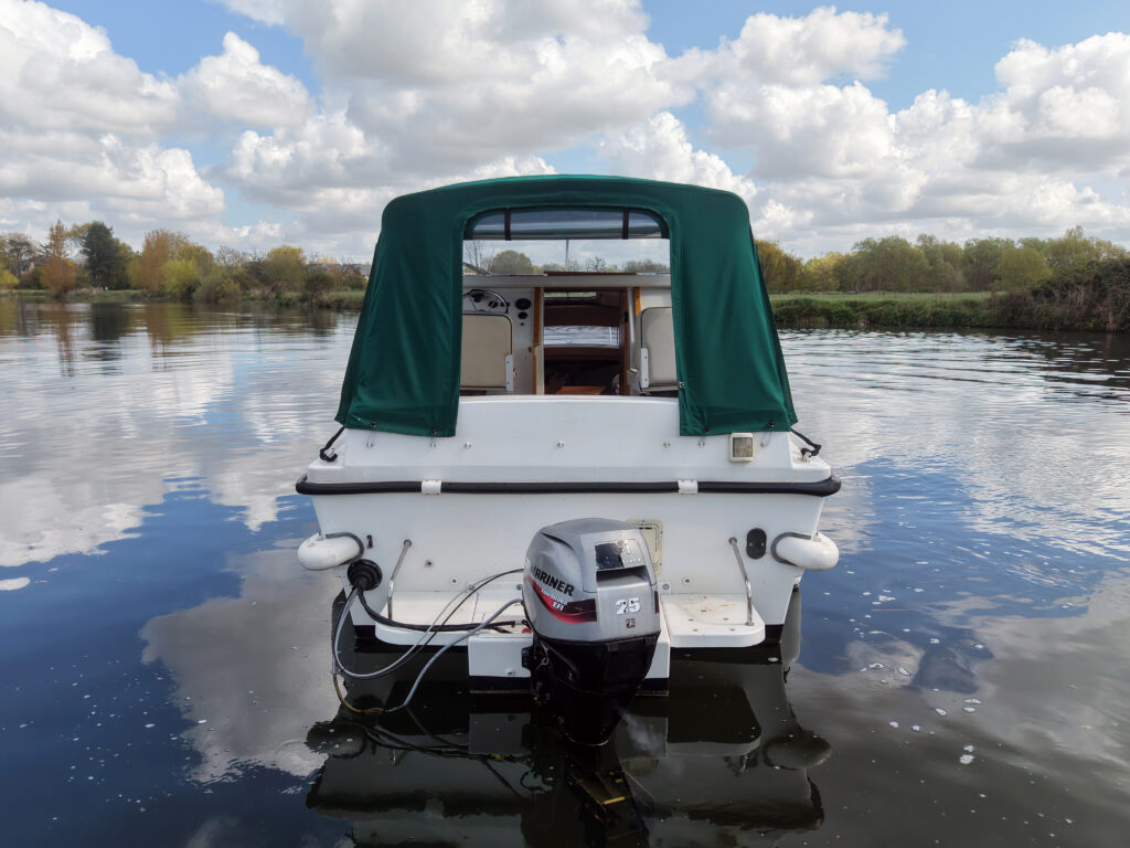 A small white boat with a green canvas cover is anchored on calm water. The boat features an outboard motor at the rear and a spacious cabin visible through the cover, while the background includes trees and blue skies with fluffy clouds.