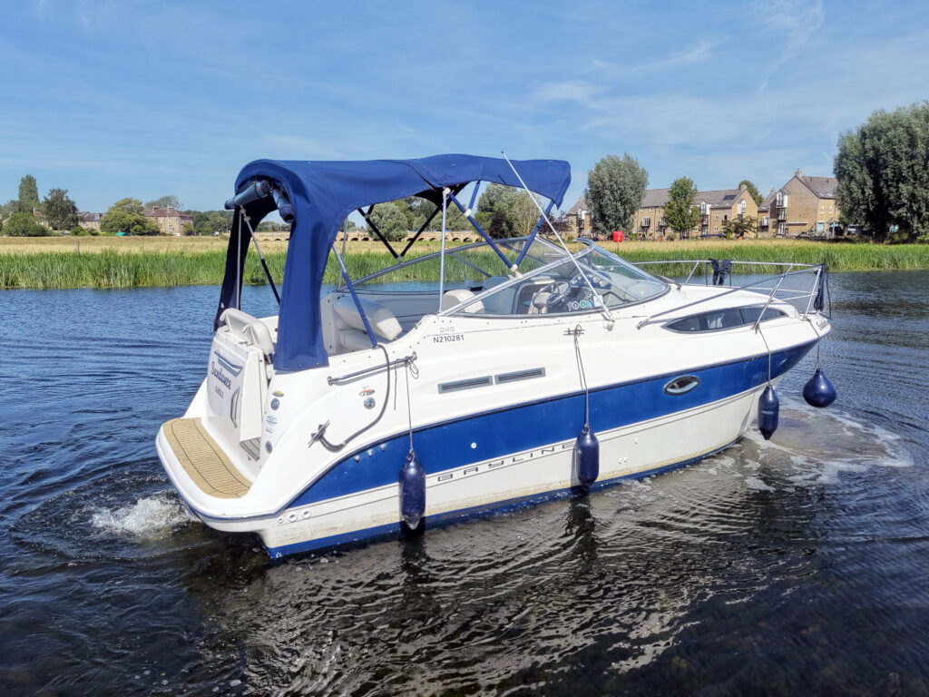 A white and blue motorboat with a navy canopy is docked on calm water. Surrounding the boat are lush green trees, and a few houses are visible in the background under a clear sky.