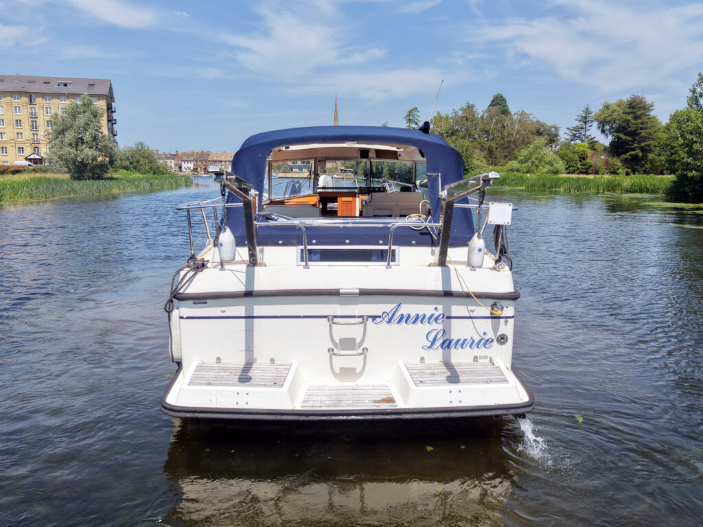 A white boat named "Annie Laurie" is moored in a calm waterway surrounded by greenery. The boat's rear view shows a blue canopy and seating area, with buildings and trees in the background under a clear blue sky.
