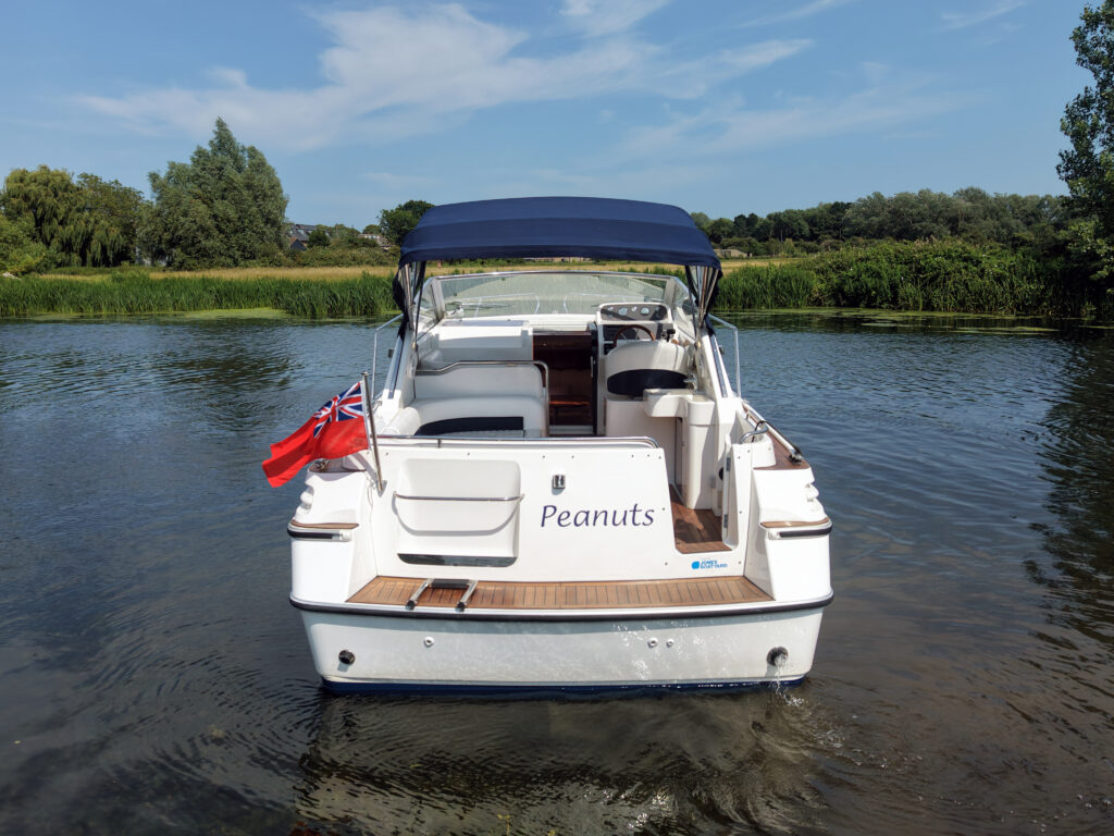 A white motorboat named "Peanuts" anchored on calm water, with a blue canopy on top. The boat displays a red flag on the left side, and green trees line the banks in the background under a clear blue sky.