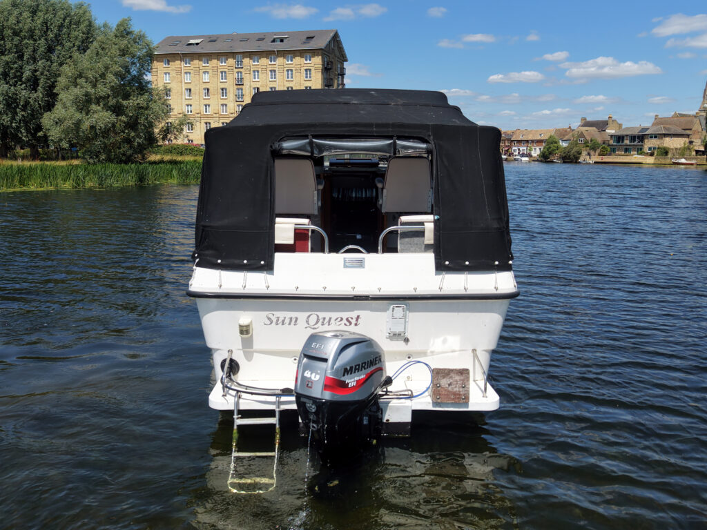 A white motorboat named 'Sun Quest' anchored on a river. The boat features a black canopy and has a motor labeled 'Mercury' attached at the stern. In the background, a brick building with multiple windows stands next to lush greenery and blue sky.