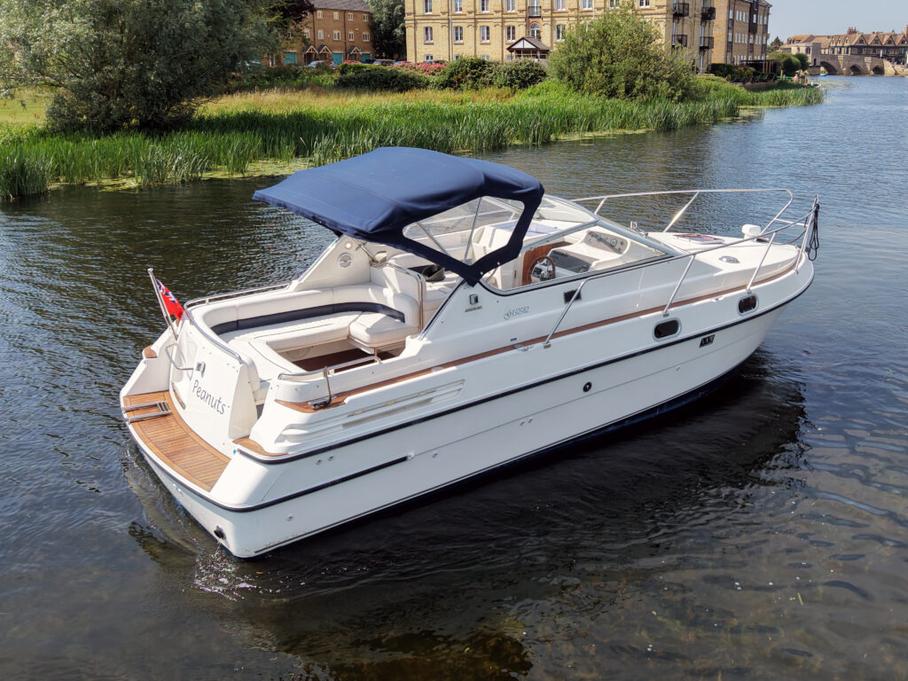 A white cabin cruiser boat with a blue canopy is anchored in a calm river. The boat displays a wooden deck and is surrounded by green foliage and buildings in the background.
