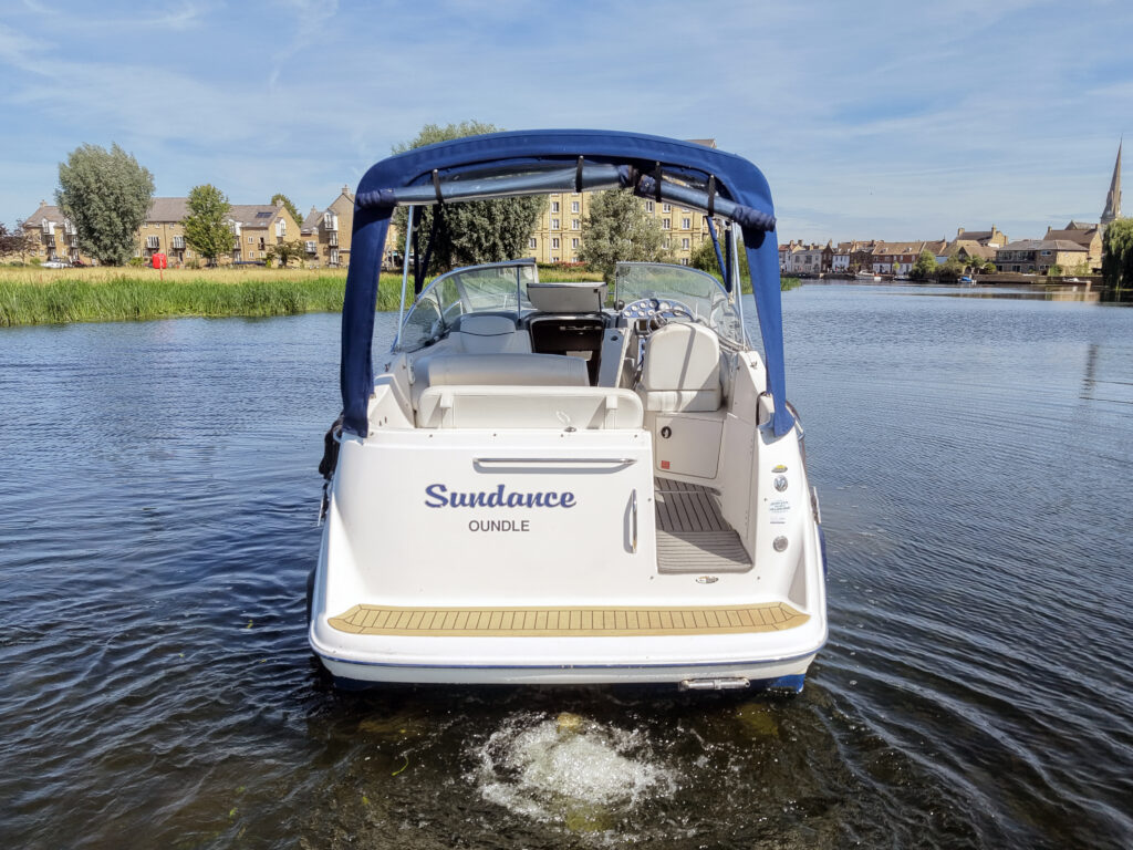 A white boat named "Sundance" is docked in a calm river, facing towards the camera. The boat features a blue canopy and is surrounded by greenery and buildings visible in the background under a clear blue sky.