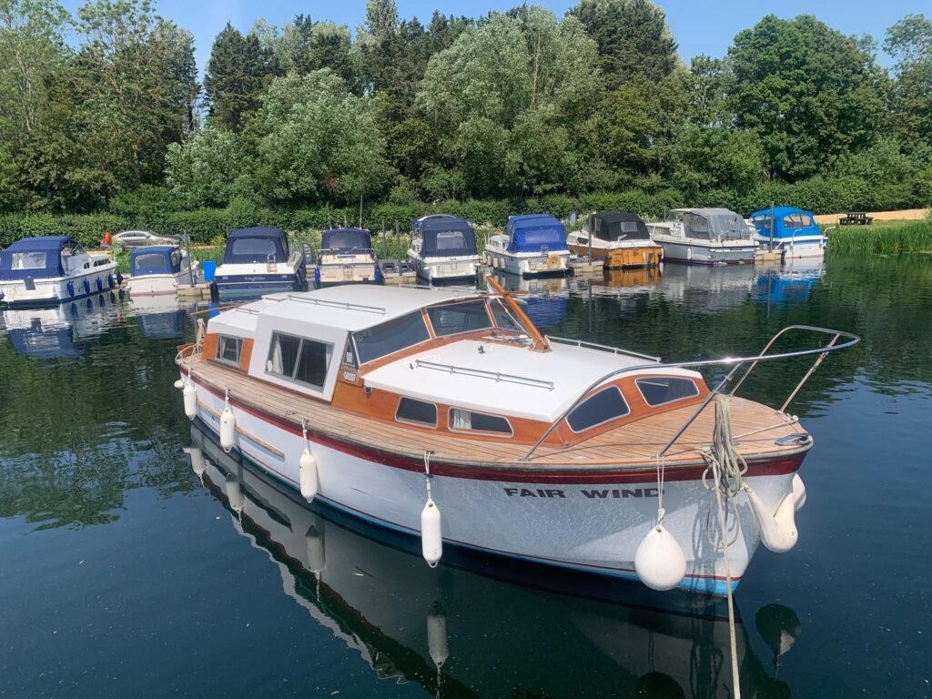 A classic L H Jones cruiser named 'Fair Winds' is moored in calm waters, featuring a wooden deck and a white hull with a red stripe. Several other boats are visible in the background, with greenery lining the shoreline.