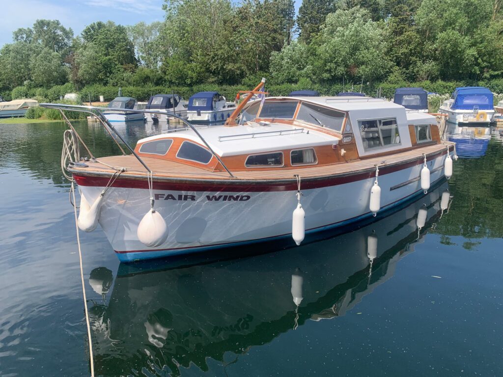 A wooden boat named "FAIR WIND" is docked on calm water, reflecting the surrounding trees and other boats in the background. The boat features a classic design with a polished wooden deck and white hull, complemented by fenders hanging off the side.