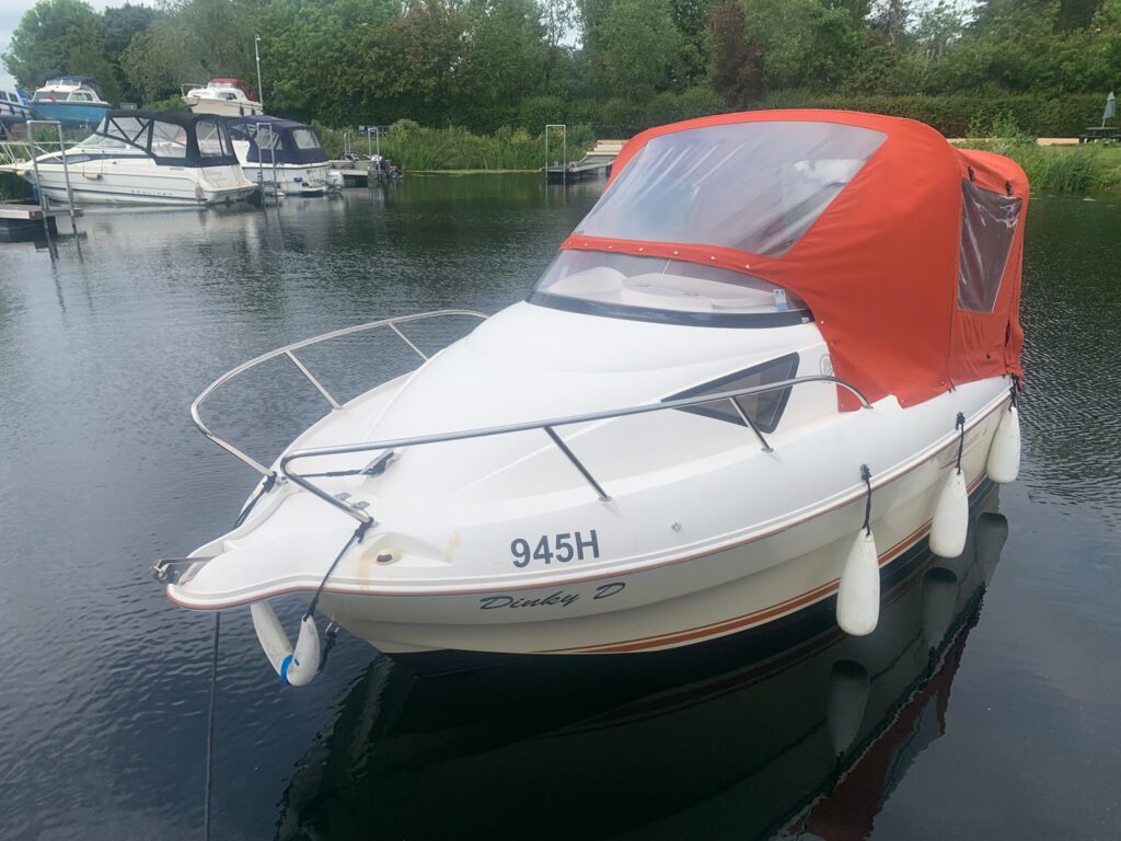 A white boat named "Dandy D" with an orange canopy is docked in calm water. The boat is equipped with fenders on the side and displays the registration number 945H. Nearby, other boats are visible in the background.