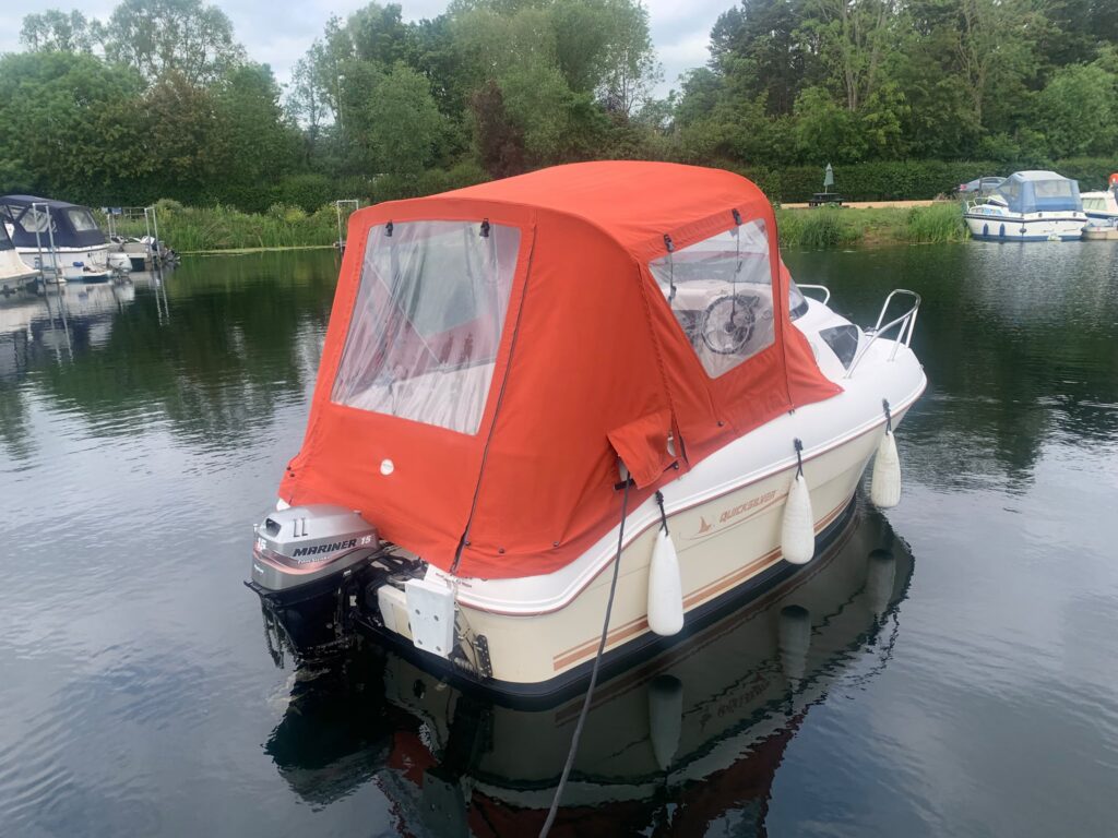 A boat docked on calm water, covered with a bright orange canopy. The boat has a white hull and several fenders hanging over the side. Surrounding greenery is visible in the background, with other boats moored nearby.