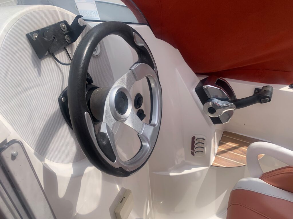 A close-up view of a boat's control panel featuring a metallic steering wheel and a throttle control lever on the right. The background includes a beige seat and a light-colored dashboard with a console.