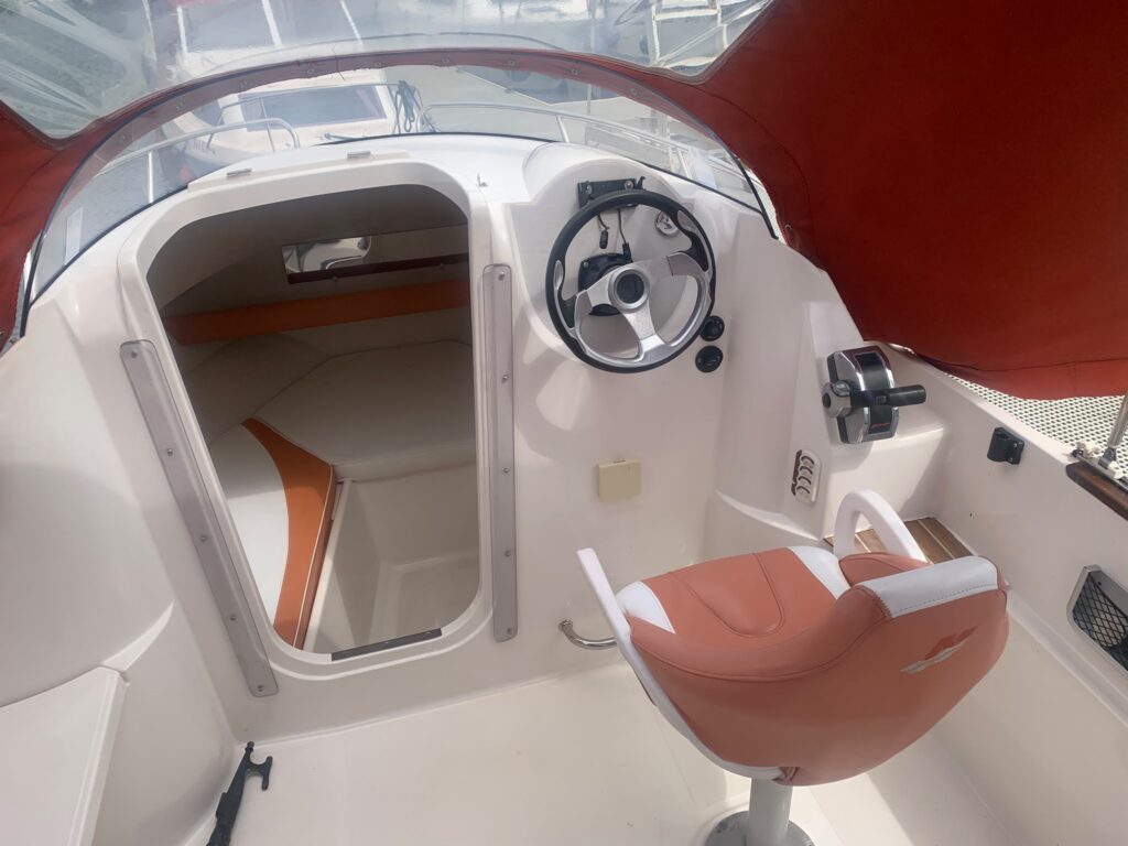 The interior of a Quicksilver 510 boat, featuring a steering wheel, a white seat with orange accents, and a partially open hatch revealing the cabin. The walls and flooring are white, with a clear canopy allowing natural light in.