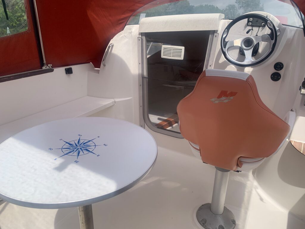 The interior of a boat features a round table with a nautical compass design, accompanied by a light brown captain's chair. A steering wheel can be seen on the dashboard, with a red canopy overhead and white surfaces surrounding the seating area.