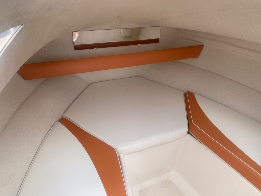 Interior view of a boat featuring a white seating area with orange accents. The seating is arranged in a hexagonal shape with a flat surface, and a small mirror is mounted above. The walls and ceiling are upholstered with beige material.
