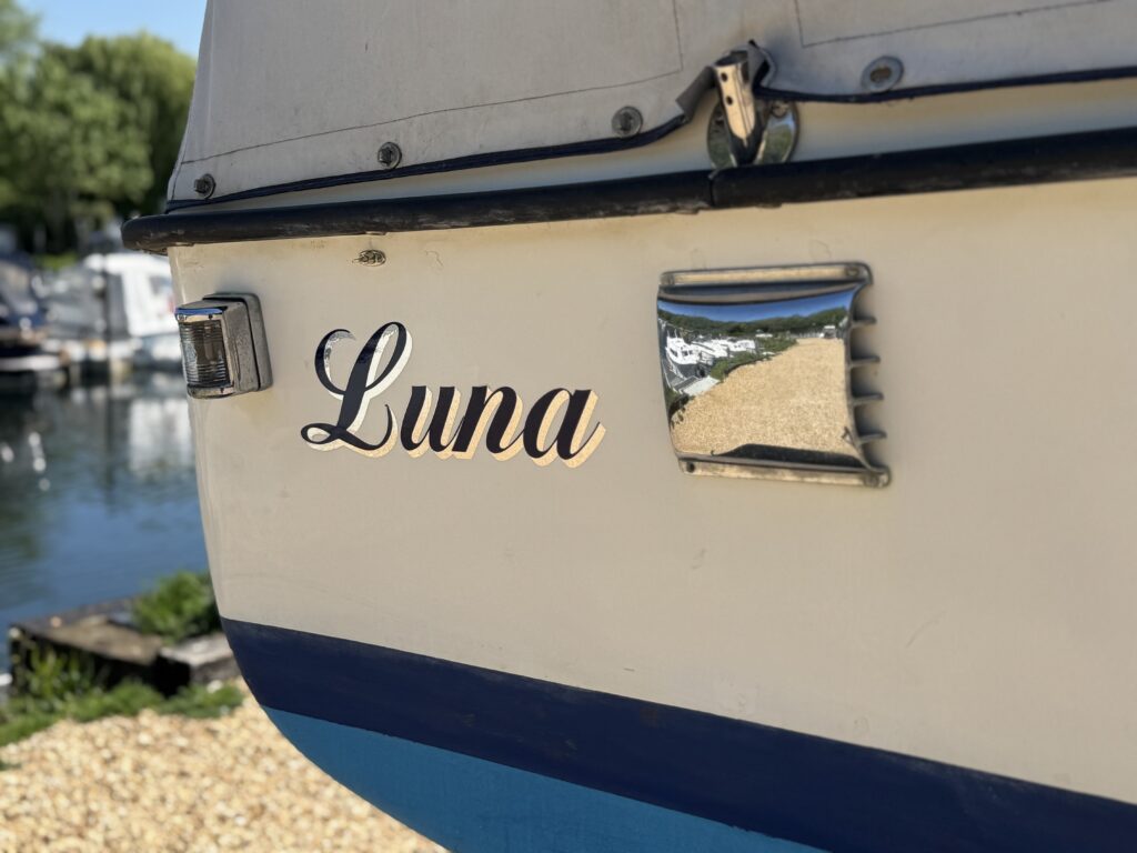 The rear of a boat displays the name "Luna" in stylish script. A chrome light fixture is positioned next to the name, reflecting the surroundings, while part of a marina is visible in the background.