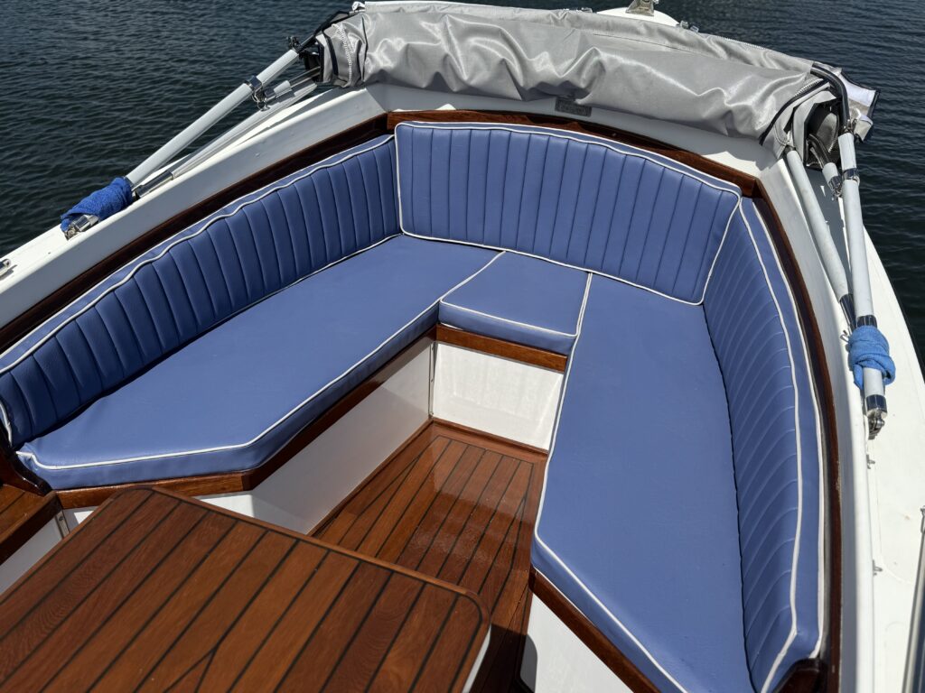 A close-up view of a boat's seating area featuring blue cushioned benches with vertical stitching and white piping. The benches are arranged around a wooden table, with a canopy overhead and water visible in the background.