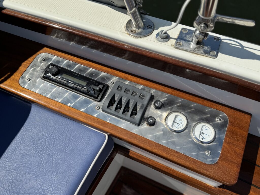 A close-up view of a boat control panel featuring a silver metal surface with various knobs and switches, including a radio. Two round gauges for measuring, possibly temperature or pressure, are positioned on the right side. The background shows part of a blue cushioned seat.