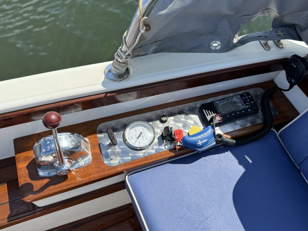 A close-up of a boat's control panel featuring a chrome throttle lever, a round pressure gauge, and a mounted speaker cable. The panel is made of polished wood and aluminum, with a blue cushioned seat visible in the foreground.