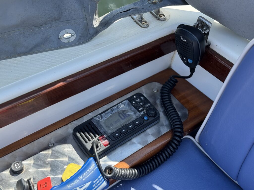 A boat's cockpit features a VHF radio with a microphone and a display showing the channel number. The radio is mounted on a wooden surface beside a blue cushioned seat, with a metal panel and equipment visible.