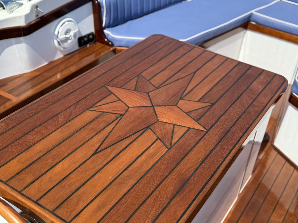 A wooden table on a boat, featuring a star inlay design. The table has a polished finish with horizontal wood planks, and the surrounding area includes blue upholstery and wooden paneling.