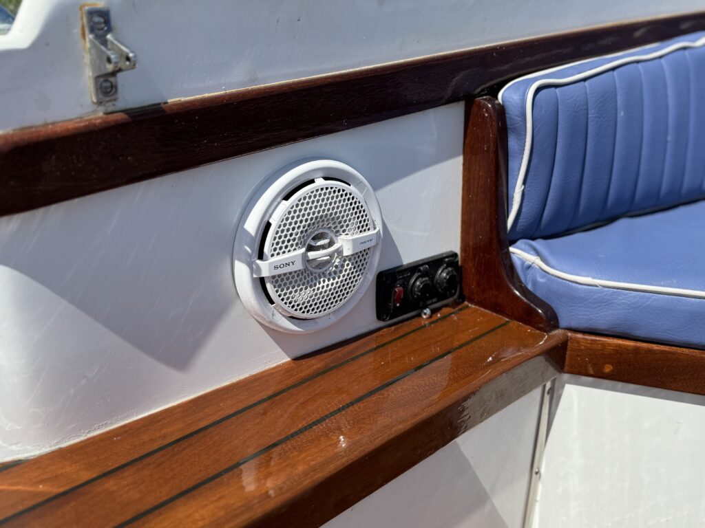 A white outdoor speaker mounted on a wooden panel in a boat, next to a control panel with two switches. A portion of blue cushioned seating is visible in the background, along with a clean white surface.