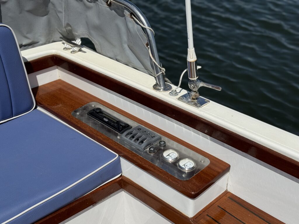 A close-up view of a boat's control panel featuring various buttons and switches on a metallic plate. The panel is mounted on a polished wooden surface next to a blue cushioned seating area, with a blurred water surface visible in the background.