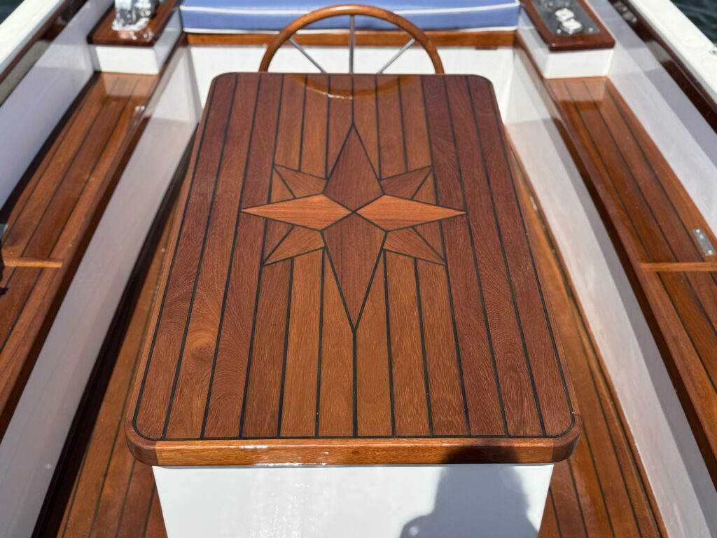 A wooden boat table with a polished finish features a star inlay design at its center. Surrounding the table are smooth wooden benches, and the boat’s interior is bright and airy with shafts of daylight illuminating the space.