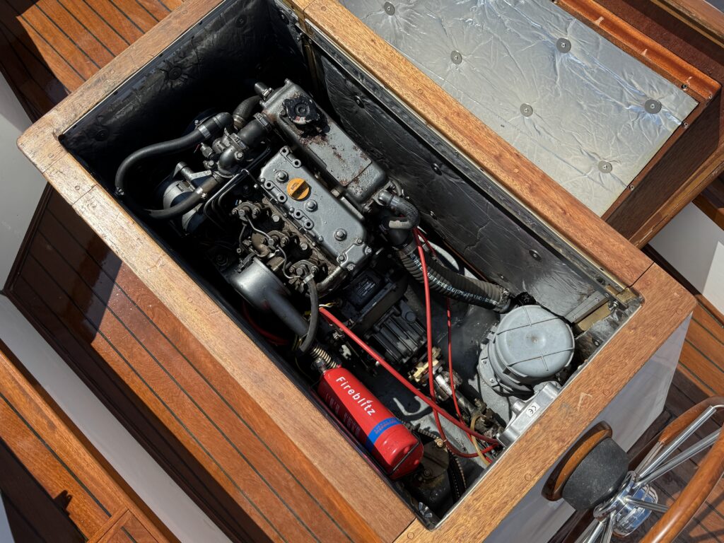 An engine compartment installed in a wooden boat, featuring various mechanical components, including a silver engine, red and black cables, and a red fire extinguisher. The lid is open, revealing the intricate setup inside.