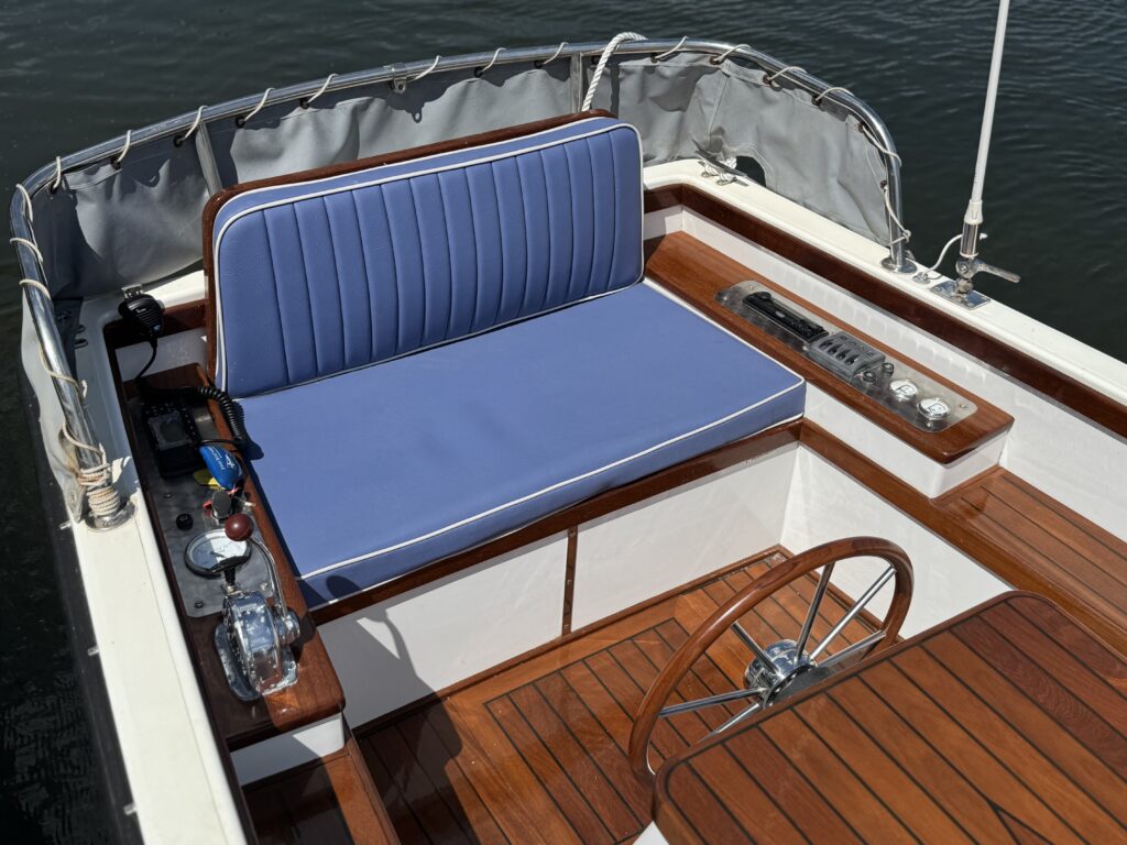 An interior view of a boat showing a blue cushioned seat with vertical stripes, situated against a wooden panel. Nearby is a steering wheel made of polished wood, a control panel with various instruments, and a smooth wooden floor.