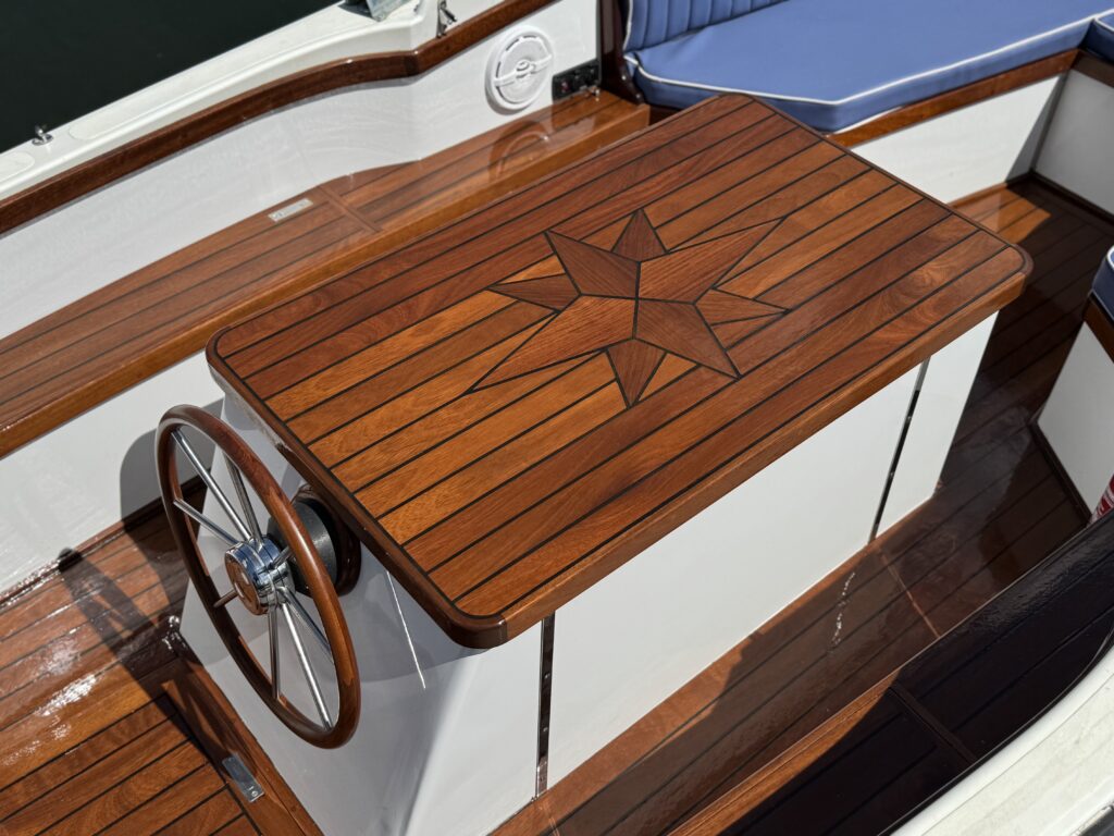 A polished wooden table with a star inlay design sits in a boat cabin. The table is surrounded by shiny wooden surfaces and a blue cushioned seat in the background, with sunlight reflecting off the smooth surfaces.