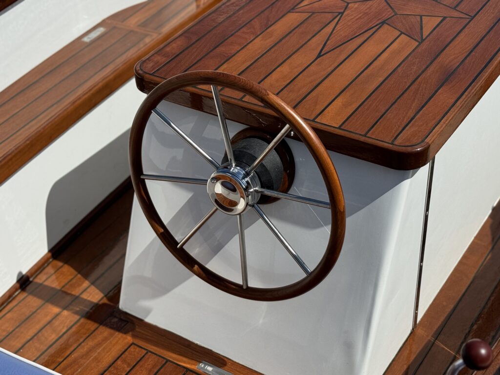 A wooden boat's helm featuring a round steering wheel, set against a polished white console. The tabletop displays a geometric pattern in rich brown wood, creating an elegant nautical aesthetic.