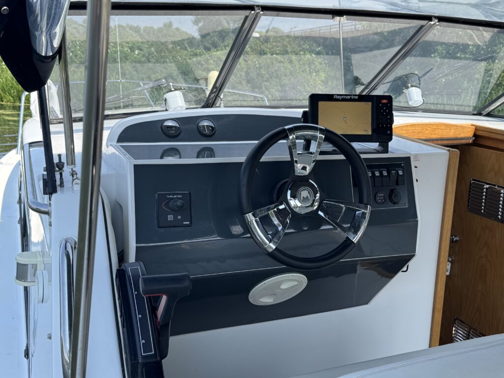 A close-up view of a boat's steering console featuring a black and gray dashboard. The console includes a steering wheel, various controls, and a digital display mounted on a panel. The background shows a clear view of greenery through the boat's windows.