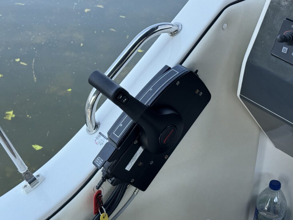 A close-up view of a boat's throttle control mounted on the side of the cockpit. The control features a black handle and several buttons, with a stainless steel railing visible in the background along with a bottle of water.
