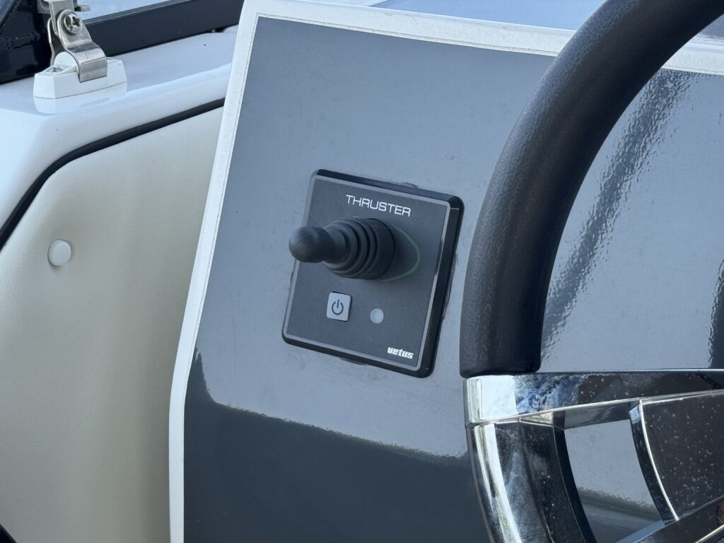 A close-up view of a boat control panel featuring a thruster joystick with a button labeled "THRUSTER" and an on/off indicator. The panel has a glossy dark surface and is positioned near a steering wheel.