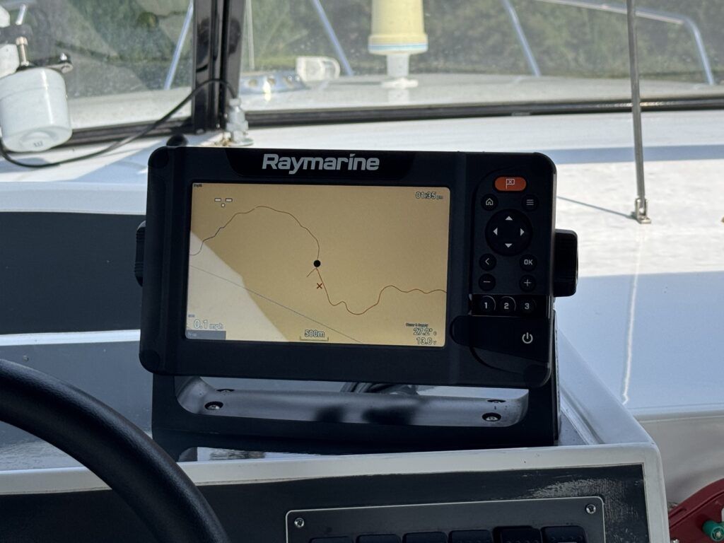 A Raymarine chart plotter display showing a map with a marked location in the center of the screen. The device is mounted in a boat dashboard with various controls visible below it.