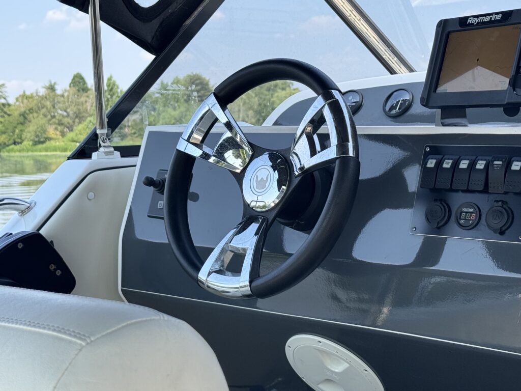 A close-up view of a chrome and black boat steering wheel mounted on a gray dashboard. The dashboard features a digital display and several control switches, with a blurred natural landscape visible in the background.
