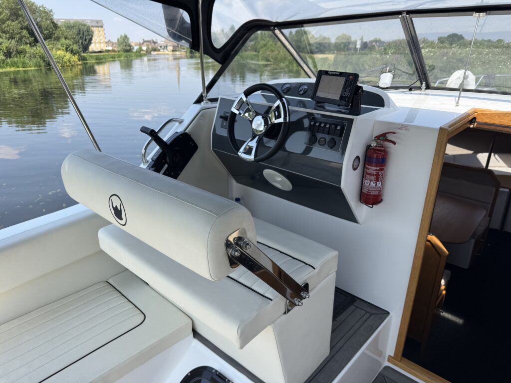The interior of a boat features a white and gray helm with a steering wheel, digital display, and various controls. A cushioned bench seat with armrests and a safety fire extinguisher is visible, alongside the calm water reflecting greenery outside.
