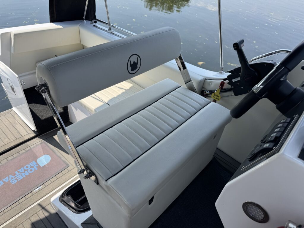 A gray padded captain's seat on a boat, featuring vertical stripes and a logo on the backrest. The seat is positioned near the boat's controls, with the water visible in the background and interior boat fixtures surrounding the area.