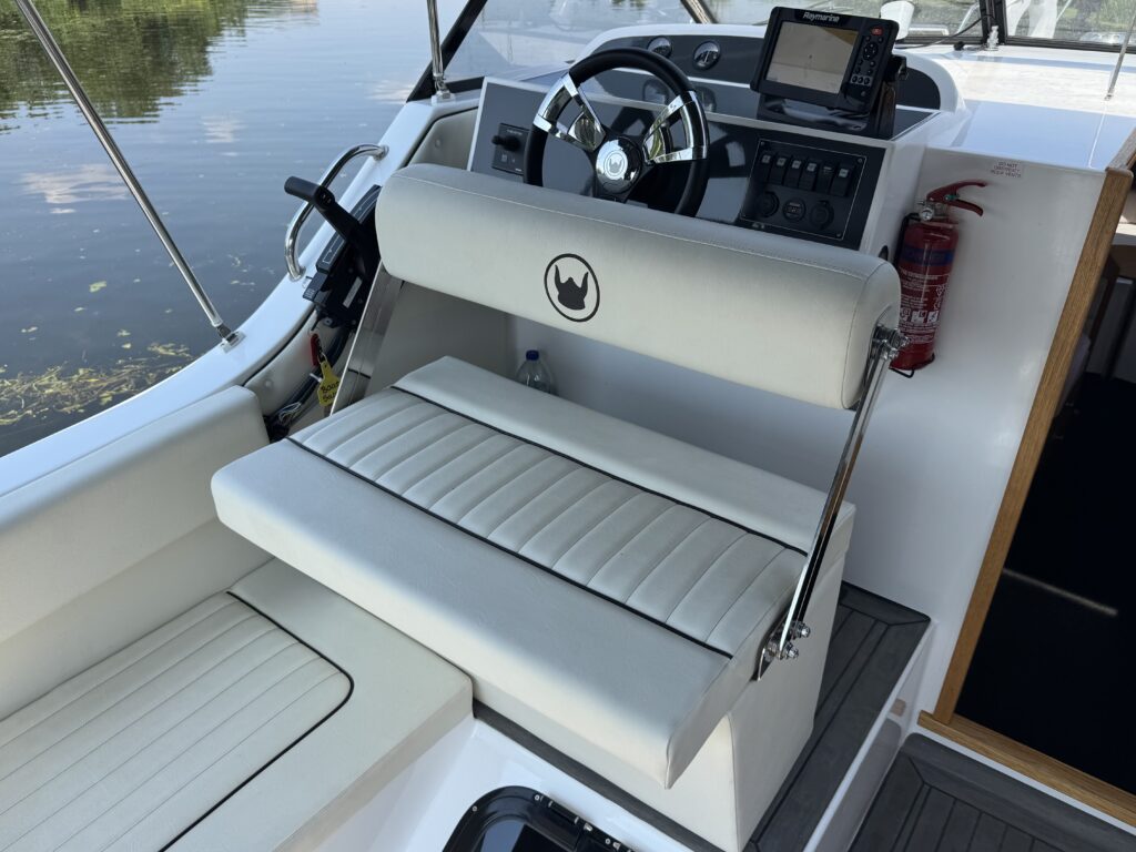 A white padded boat seat with horizontal stitching is positioned on the deck of a boat. The steering wheel and a navigation display are visible in the background, along with a fire extinguisher mounted on the side.