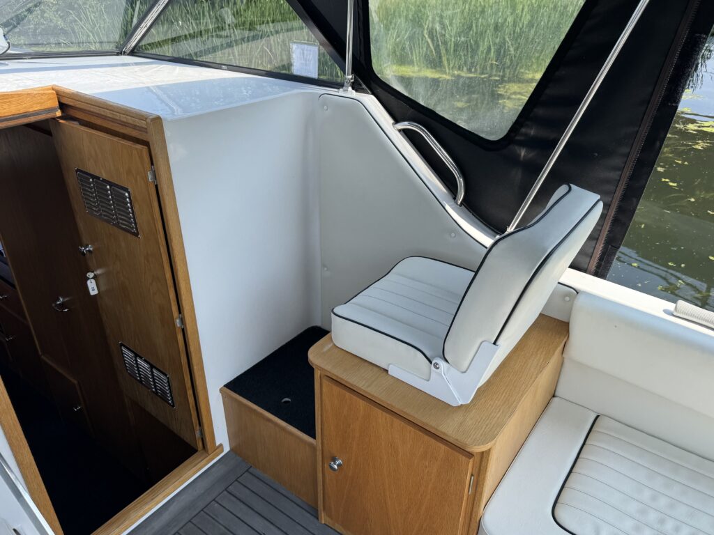 An interior view of a boat featuring a wooden cabinet with metal mesh vents, a white cushioned seat on a wooden pedestal, and an additional white bench on one side. The background includes tall green grass visible through the windows.