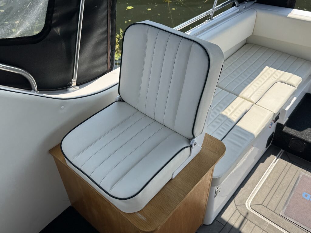 A white cushioned seat with vertical stitching is positioned on a wooden base in a boat. The background features additional beige seating and large windows showing natural light, with greenery visible outside.