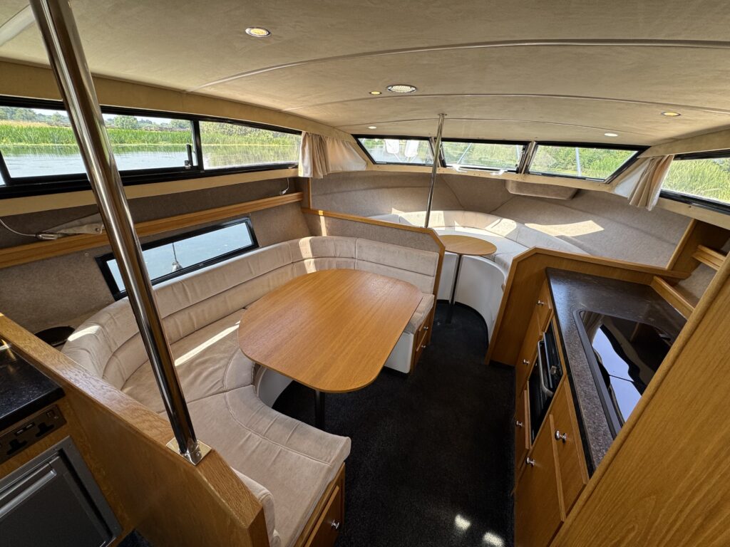 Interior view of a yacht cabin featuring a circular dining area with a wooden table surrounded by a curved cushioned bench. The kitchen area includes a countertop and cabinets, with large windows providing natural light and views of greenery outside.