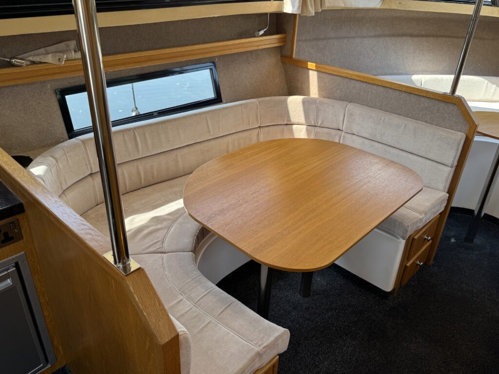 An interior view of a boat featuring a U-shaped seating area with beige cushions around a wooden table. A window provides natural light, and the floor is covered with dark carpet.