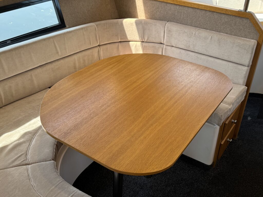 A wooden table with a smooth surface is positioned at the center of a curved beige sofa. The sofa features padded seats, and natural light streams in through a nearby window, highlighting the furniture.