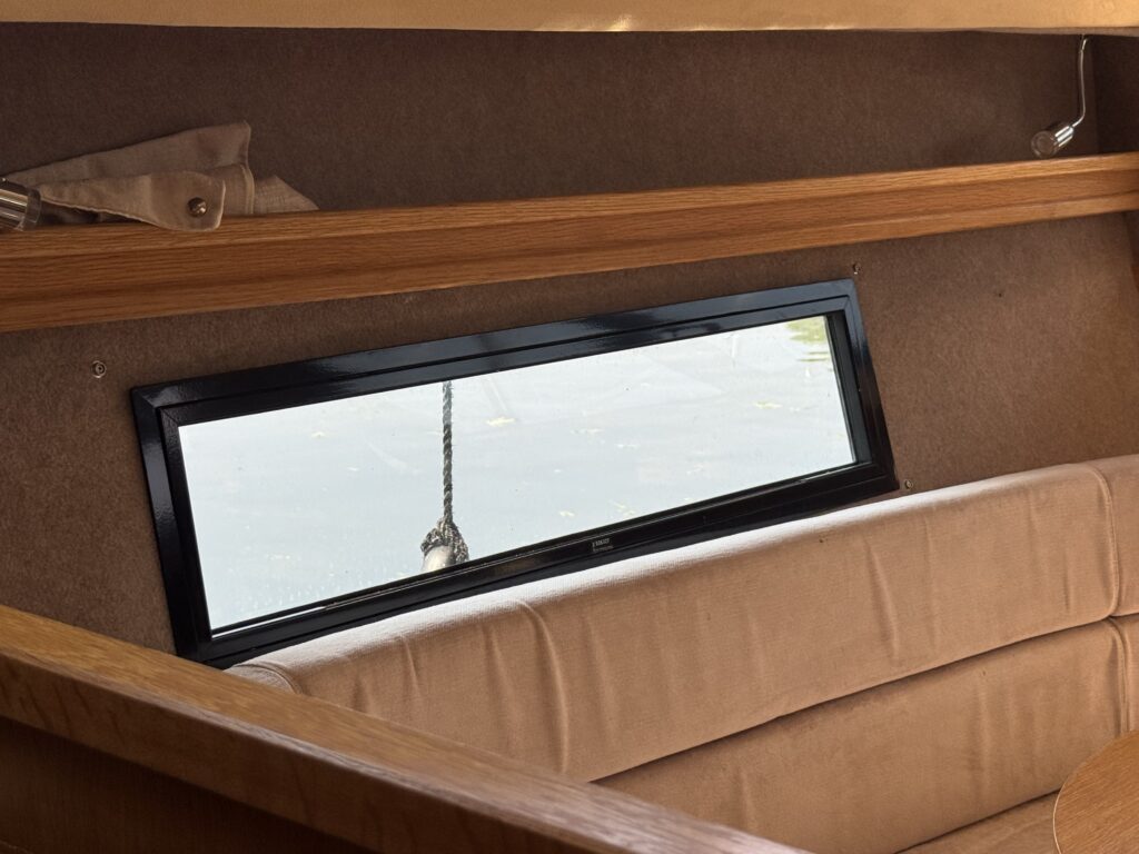 An interior view of a boat showing a brown leather bench seat facing a horizontal window. The window has a black frame and provides a view of water outside, with a rope hanging near it. The wall above has a wooden shelf.