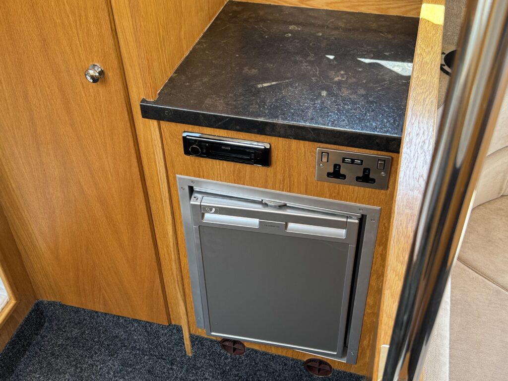 A wooden cabinet featuring a black countertop and a silver-colored refrigerator with a pull-down door. Above the refrigerator, there is a car radio and two electrical outlets mounted on the wall near the cabinet.
